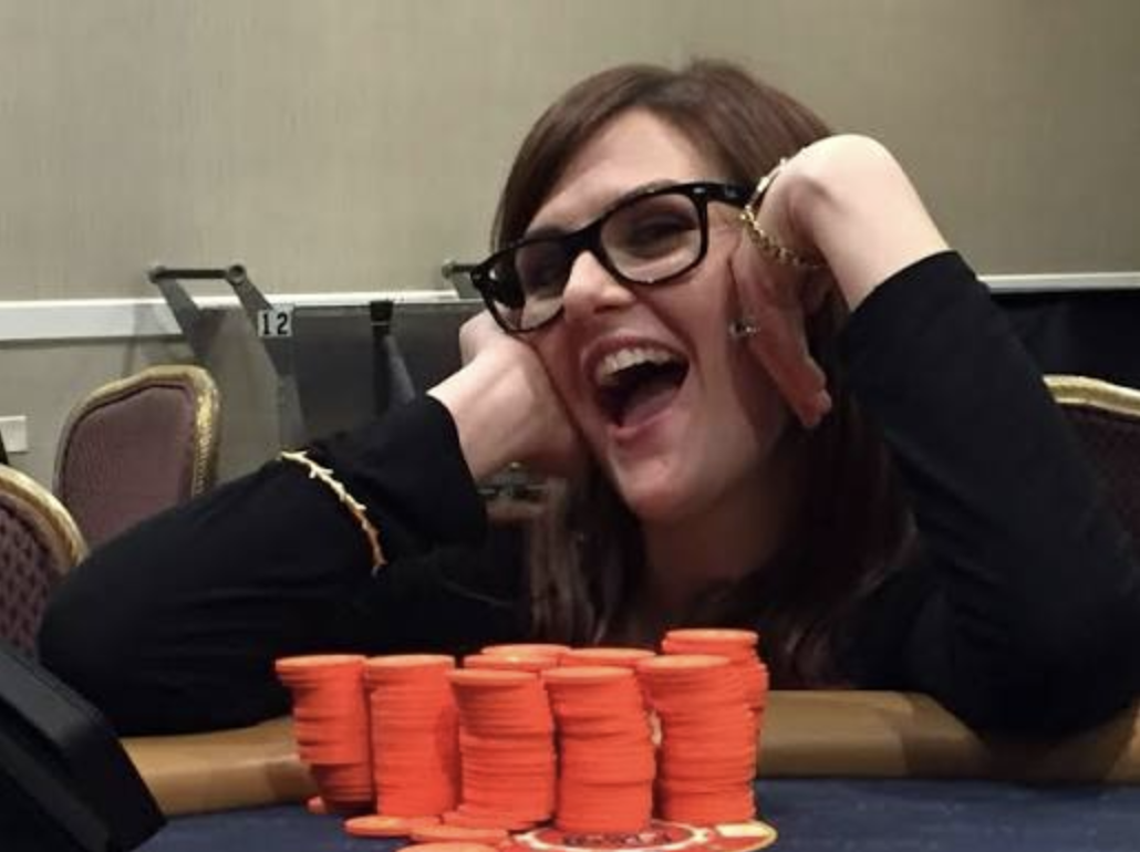 Sara Rue in glasses leans on her elbow behind tall stacks of orange tournament chips.