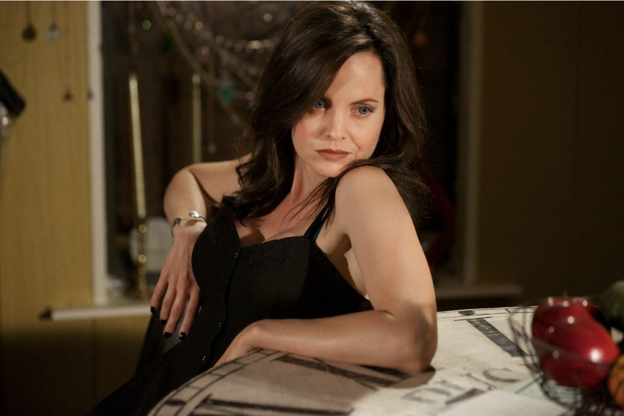 Mena Suvari in a black dress poses at a kitchen table in a dim, string-lit room.