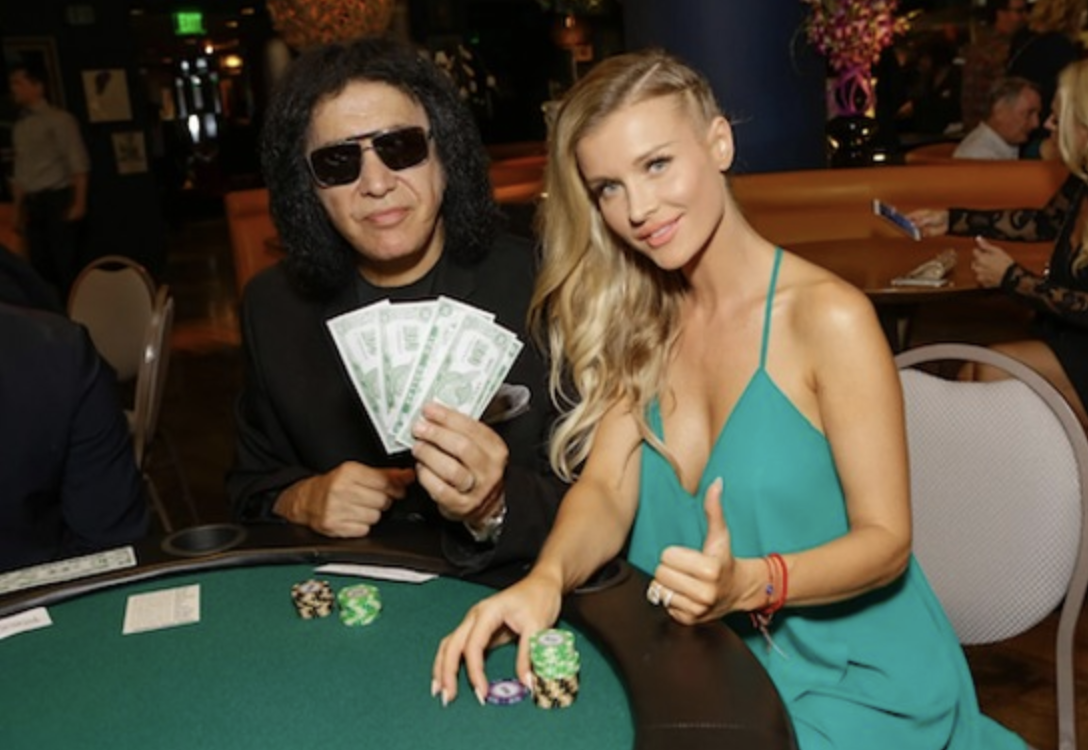 Rock musician Gene Simmons fans out novelty cash beside Joanna Krupa giving a thumbs-up at a charity poker table.